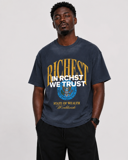 A man wearing a dark navy graphic t-shirt with a distressed print that reads 'RICHEST IN RCHST WE TRUST' in gold and white, with a blue circular emblem below. He is also wearing black pants, standing against a light gray studio background.