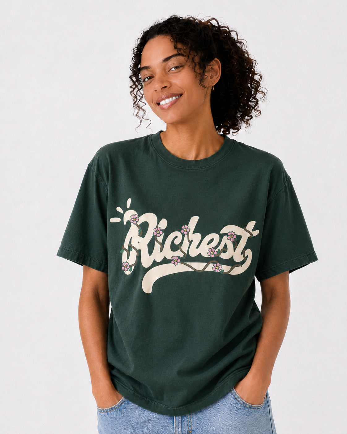 A woman wearing a dark green graphic t-shirt with the word 'Richest' in a retro cream font decorated with small purple flowers, paired with light blue jeans, against a plain light white background.